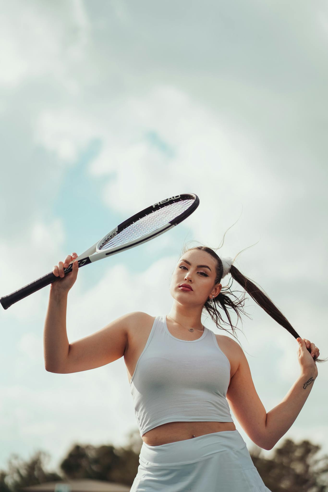 Young woman posing with a tennis racket against a cloudy sky.